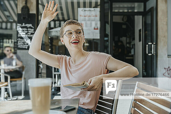 Woman with book waving while sitting in sidewalk cafe on sunny day