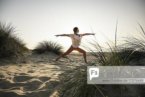 Junge Frau übt Krieger 2 Position Yoga inmitten von Pflanzen am Strand gegen klaren Himmel bei Sonnenuntergang