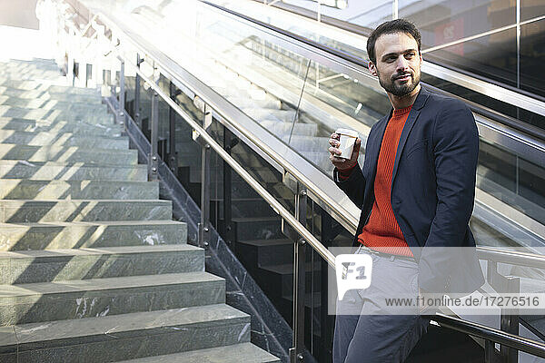 Selbstbewusster Unternehmer trinkt Kaffee  während er auf einer Treppe in der Stadt steht