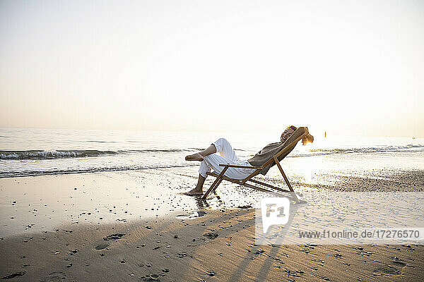Lächelnde junge Frau  die sich auf einem Klappstuhl am Strand vor einem klaren Himmel bei Sonnenuntergang entspannt