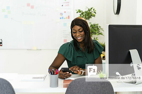 Lächelnde Frau  die ein Mobiltelefon benutzt  während sie am Schreibtisch im Büro sitzt