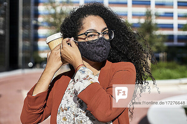 Frau trägt einen Gesichtsschutz und hält einen wiederverwendbaren Bambusbecher in der Stadt an einem sonnigen Tag