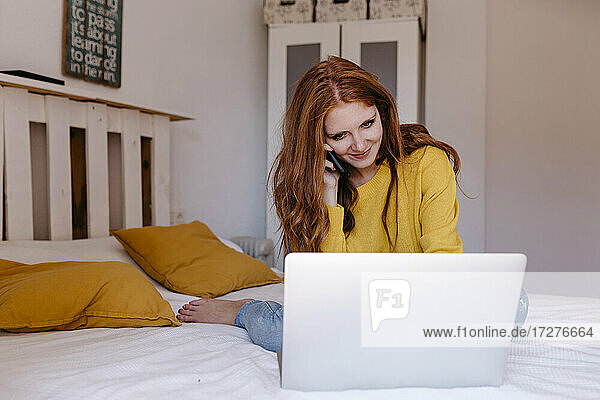 Junge Frau spricht mit ihrem Smartphone  während sie einen Laptop im Schlafzimmer zu Hause benutzt