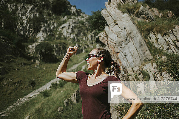Smiling woman flexing muscles while standing against rock formation on sunny day