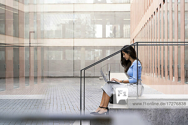 Geschäftsfrau arbeitet an einem Laptop  während sie auf den Stufen vor einem Bürogebäude sitzt