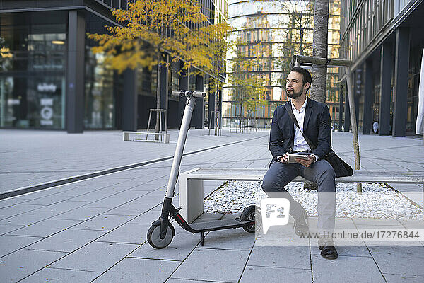 Geschäftsmann mit digitalem Tablet sitzt auf einer Bank in der Stadt neben einem E-Scooter