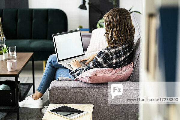 Junge Frau arbeitet am Laptop  während sie zu Hause auf dem Sofa sitzt