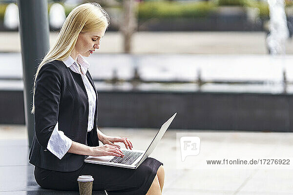 Blonde Geschäftsfrau arbeitet am Laptop  während sie auf einer Bank sitzt