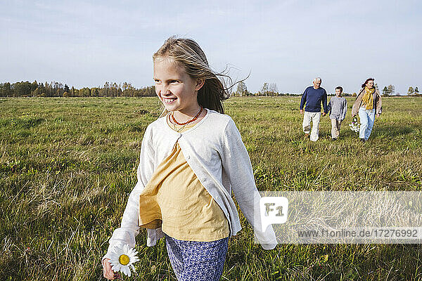 Lächelndes Mädchen  das auf einem Feld läuft  während die Familie im Hintergrund zu sehen ist