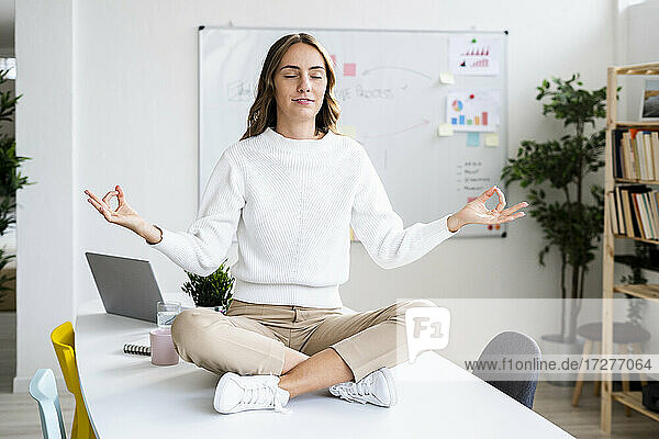 Junge Geschäftsfrau übt Yoga  während sie auf dem Tisch im Büro sitzt
