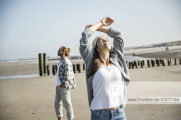 Junge Frau mit geschlossenen Augen und erhobenen Armen am Strand stehend an einem sonnigen Tag