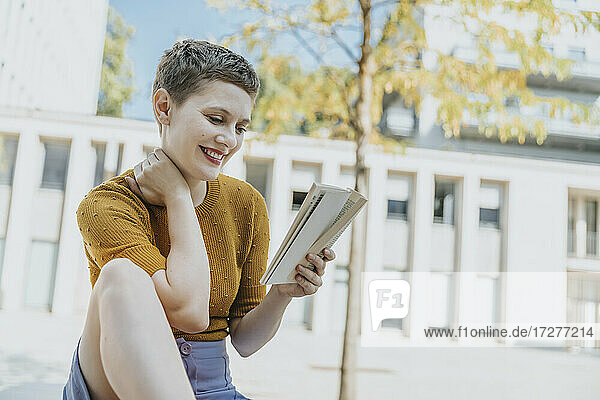 Lächelnde Frau liest ein Buch  während sie an einem sonnigen Tag auf der Straße sitzt