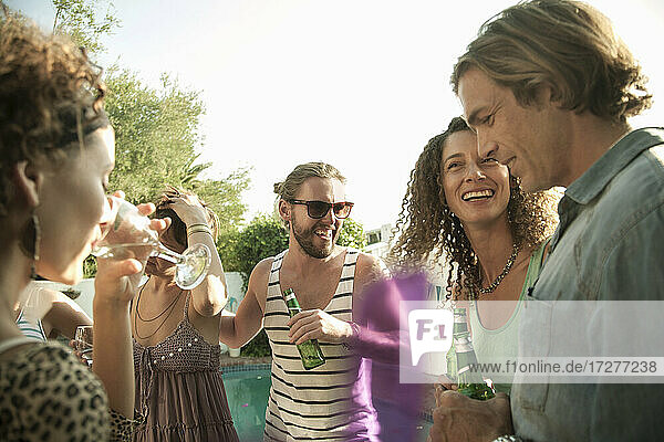 Glückliche männliche und weibliche Freunde genießen den Sommer beim Trinken von Alkohol am Pool gegen den Himmel