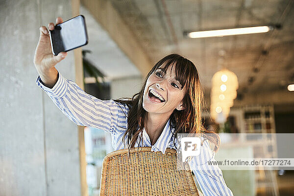 Glückliche reife Frau mit offenem Mund  die ein Selfie mit ihrem Smartphone macht  während sie zu Hause sitzt