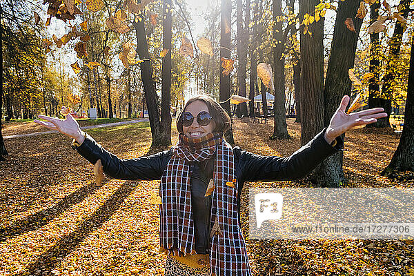 Lächelnde schöne Frau mit ausgestreckten Armen  die trockene Blätter wirft  während sie in einem Park im Herbst steht