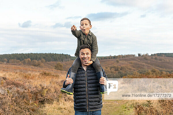 Smiling father carrying son on shoulder pointing while standing in Cannock Chase park against cloudy sky during autumn