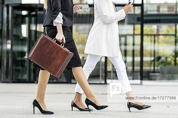 Young businesswoman with briefcase using mobile phone while walking by colleague on footpath