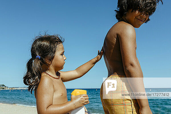 Nettes Mädchen  das Sonnencreme auf den Rücken seines Bruders aufträgt  während es einen sonnigen Tag am Strand genießt