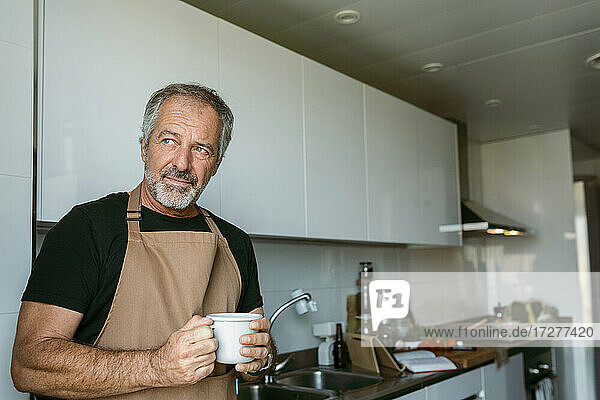 Reifer Mann  der wegschaut und eine Kaffeetasse hält  steht in der Küche zu Hause