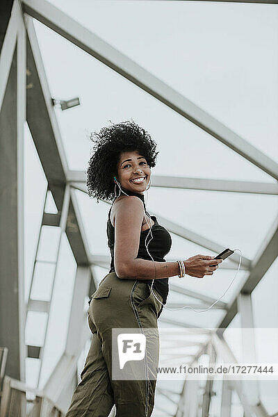 Smiling woman listening to music through earphones standing on bridge in city