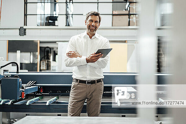 Lächelnder reifer Geschäftsmann  der ein digitales Tablet hält  während er vor einer Maschine in einer Fabrik steht