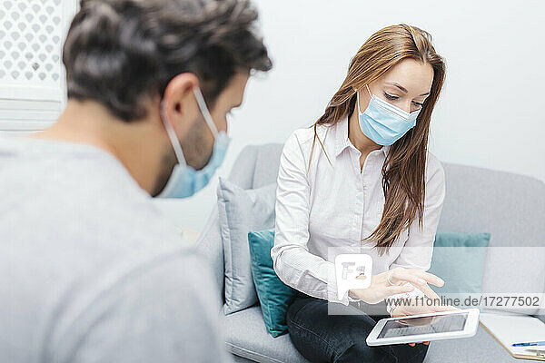 Arzt mit Gesichtsmaske  der ein digitales Tablet benutzt  während er neben einem Mann im Büro sitzt