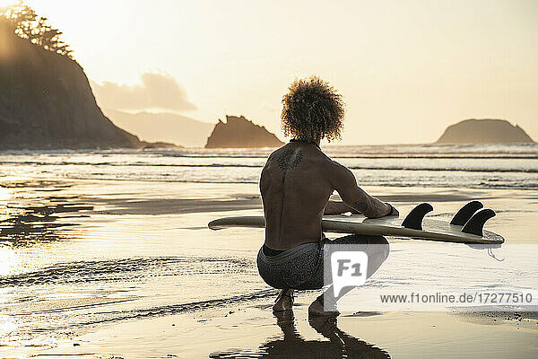 Shirtless young man with surfboard crouching at seashore during sunset