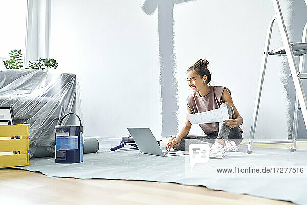 Lächelnde Frau sitzt auf dem Boden und arbeitet zu Hause am Laptop