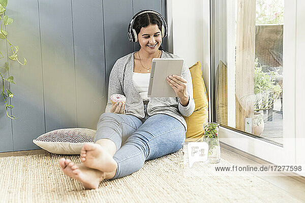 Junge Frau mit Donut  die zu Hause sitzend ein digitales Tablet benutzt