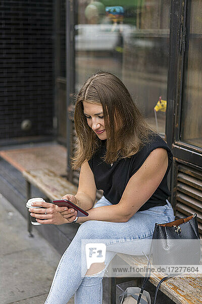Junge Frau mit Smartphone und Einweg-Kaffeebecher auf einer Bank sitzend