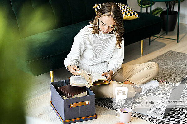 Geschäftsfrau  die ein Buch liest  während sie neben einem Laptop und einer Kiste auf dem Boden im Büro sitzt