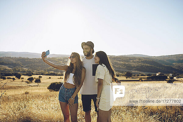 Happy male and female friends taking selfie through mobile phone while standing on field against sky