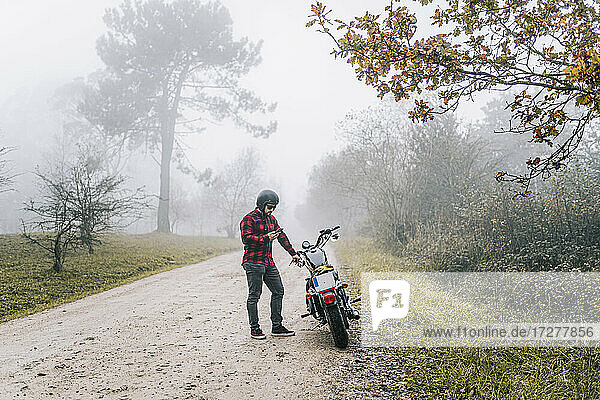 Mann benutzt sein Smartphone  während er auf einem unbefestigten Weg neben einem Motorrad steht