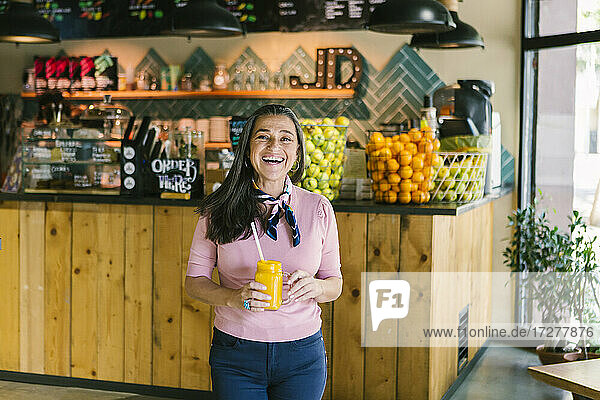 Fröhliche reife Frau  die ein Einmachglas mit frischem Saft hält  während sie in ein Café geht