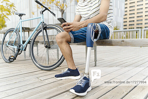 Tiefschnitt eines Mannes mit Beinprothese  der auf einer Bank in der Stadt sitzt und ein Mobiltelefon benutzt