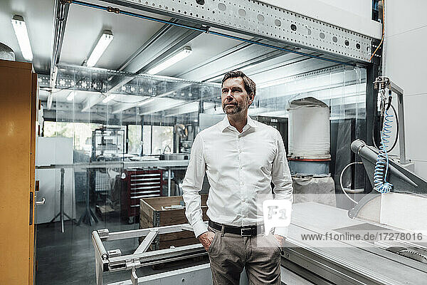 Nachdenklicher männlicher Ingenieur  der mit den Händen in den Taschen vor Maschinen in einer Fabrik steht