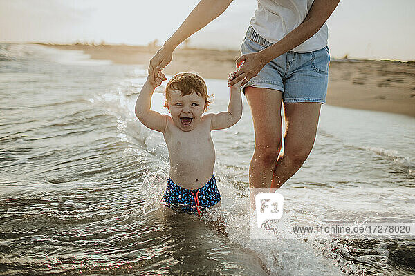 Midsection of mother with cheerful baby son enjoying in water at beach on sunny day during sunset