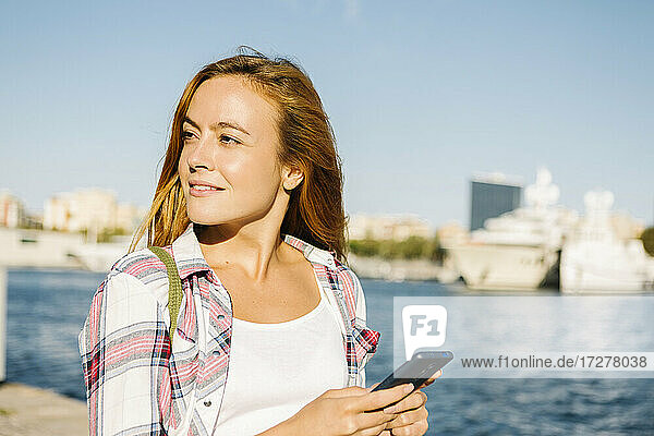 Frau benutzt Smartphone und schaut weg  während sie an einem sonnigen Tag am Meer steht