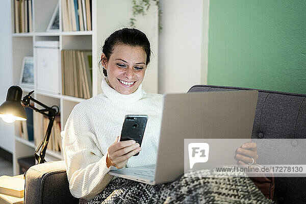 Lächelnde Frau benutzt Smartphone und Laptop  während sie zu Hause sitzt
