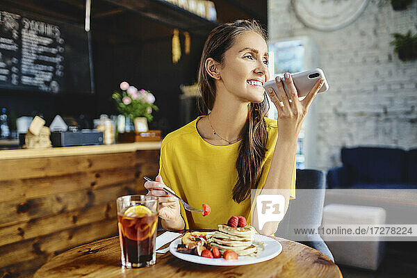 Lächelnde junge Frau isst Beeren  während sie ihr Smartphone am Tisch im Café benutzt