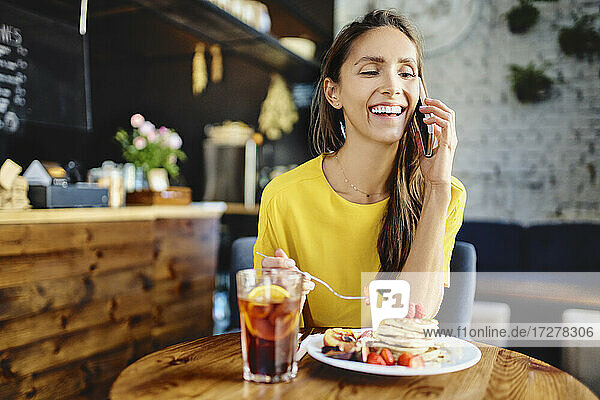 Smiling young female eating berry while talking on smart phone at table in cafe
