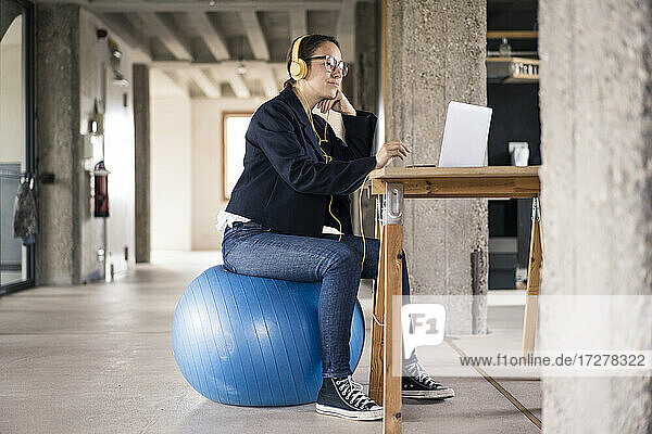 Geschäftsfrau mit Kopfhörern  die einen Laptop benutzt  während sie auf einem Fitnessball im Büro sitzt