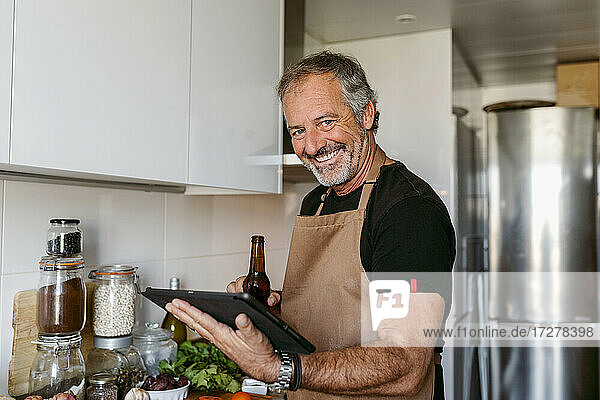 Lächelnder Mann  der eine Bierflasche und ein digitales Tablet hält  während er zu Hause in der Küche steht