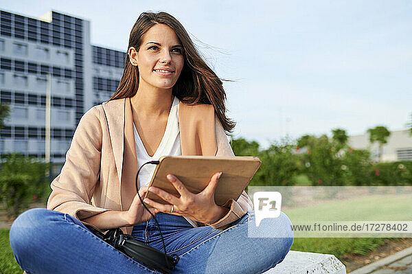 Lächelnde junge Frau  die mit einem digitalen Tablet auf einer Bank in einem öffentlichen Park sitzt und wegschaut