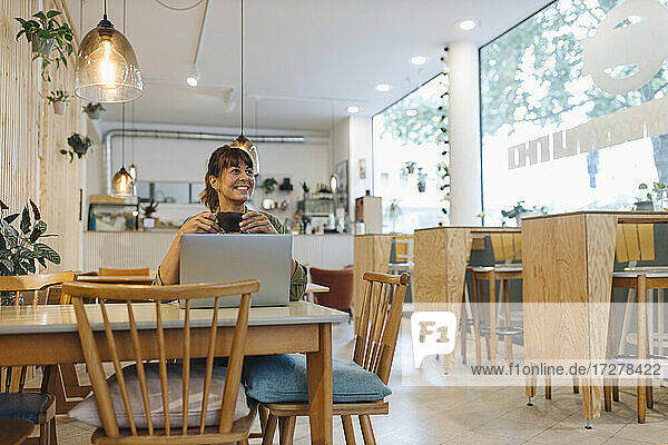 Lächelnde Unternehmerin  die eine Kaffeetasse hält und einen Laptop benutzt  während sie wegschaut und in einem Café sitzt