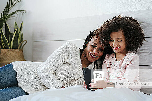 Lächelnde Mutter und Tochter mit Smartphone auf dem Bett im Schlafzimmer zu Hause sitzen