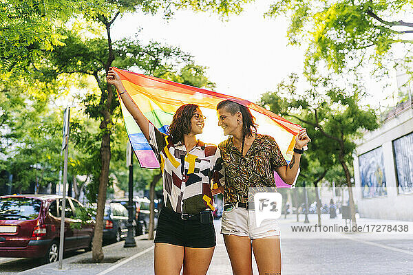 Lesbisches Paar  das sich gegenseitig anschaut  während es eine Regenbogenfahne auf einem Fußweg in der Stadt hält