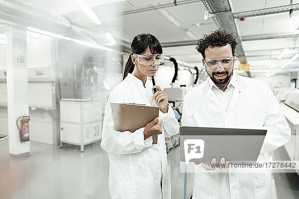 Lächelnder männlicher Wissenschaftler  der einen Laptop benutzt und neben einer Technikerin im Labor steht