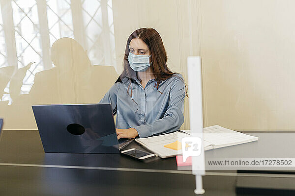 Unternehmerin mit Schutzmaske bei der Arbeit an einem Laptop am Arbeitsplatz  gesehen durch ein Fensterglas