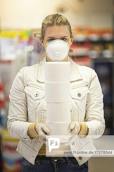 Teenager mit Schutzmaske und Handschuhen hält einen Stapel von vier Toilettenrollen im Supermarkt